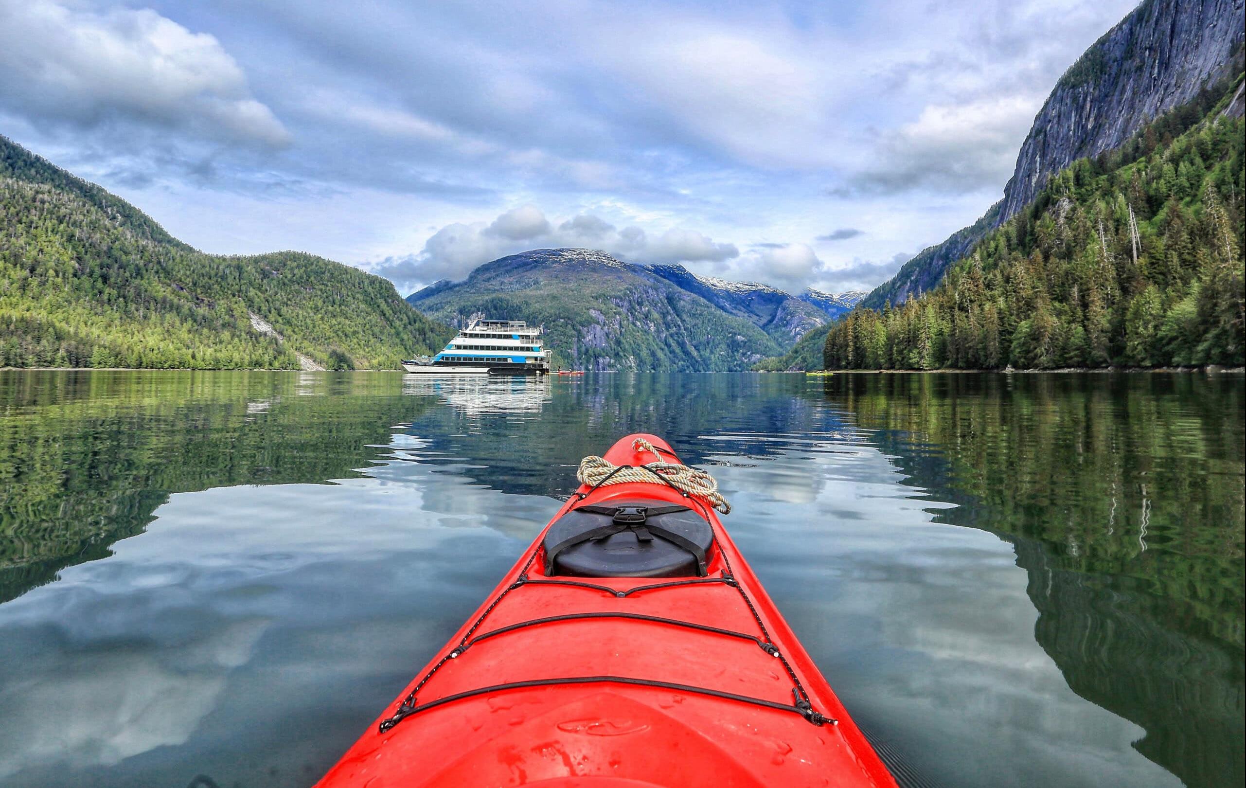 Alaska – Kreuzfahrt durch majestätische Fjorde - Die Spitze eines rote Kajaks, das durch einen Fjord mit bewaldeten Hängen fährt